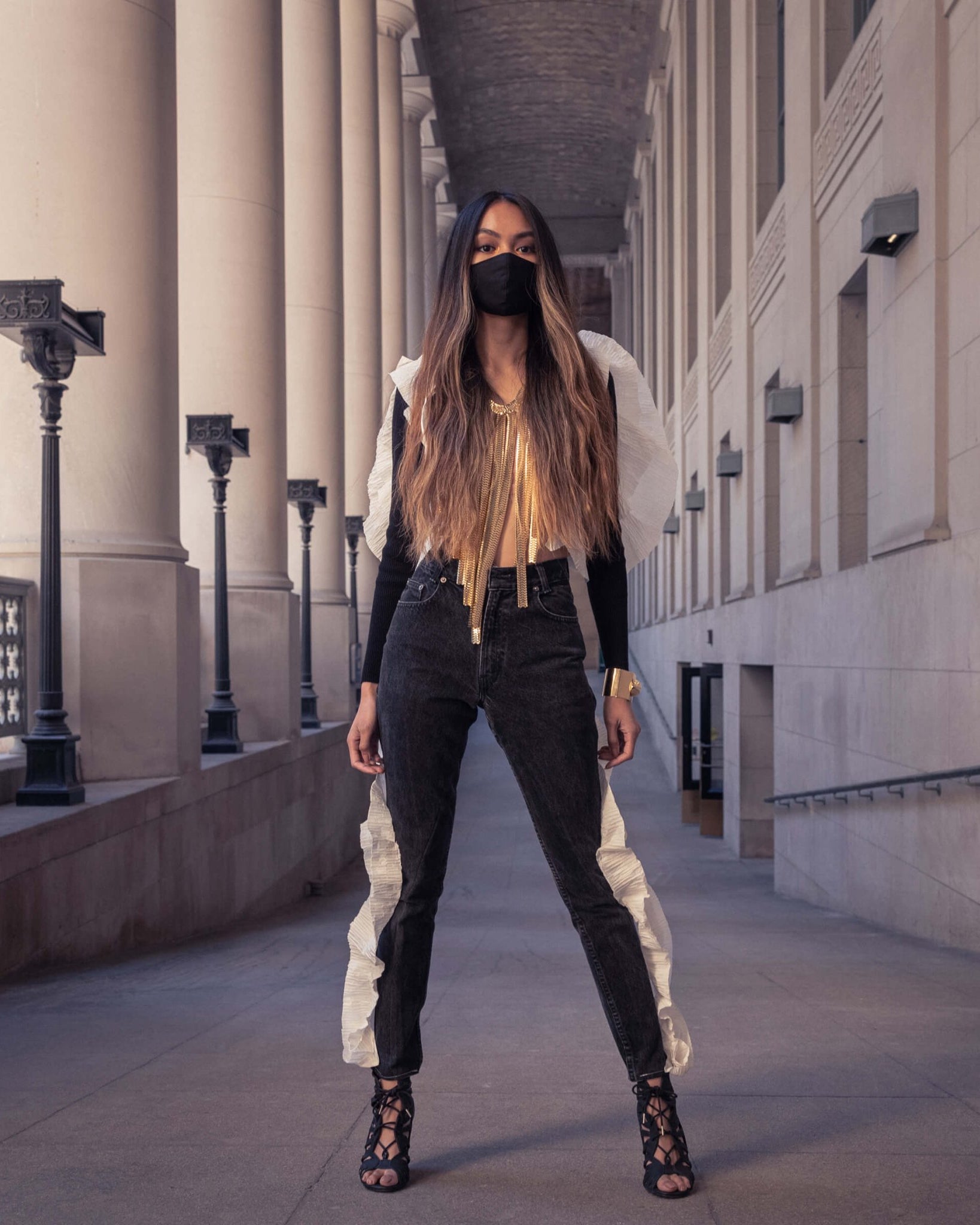 Striking model with wavy long ombré hair standing very confidently in an outdoor grand neoclassical colonnade.. She is wearing repurposed black jeans and a black bolero sweater with pleated paper ruffles sewn on to the clothes. The paper looks like wings on her back. Accented the look with a large gold fringe necklace for a dramatic look.