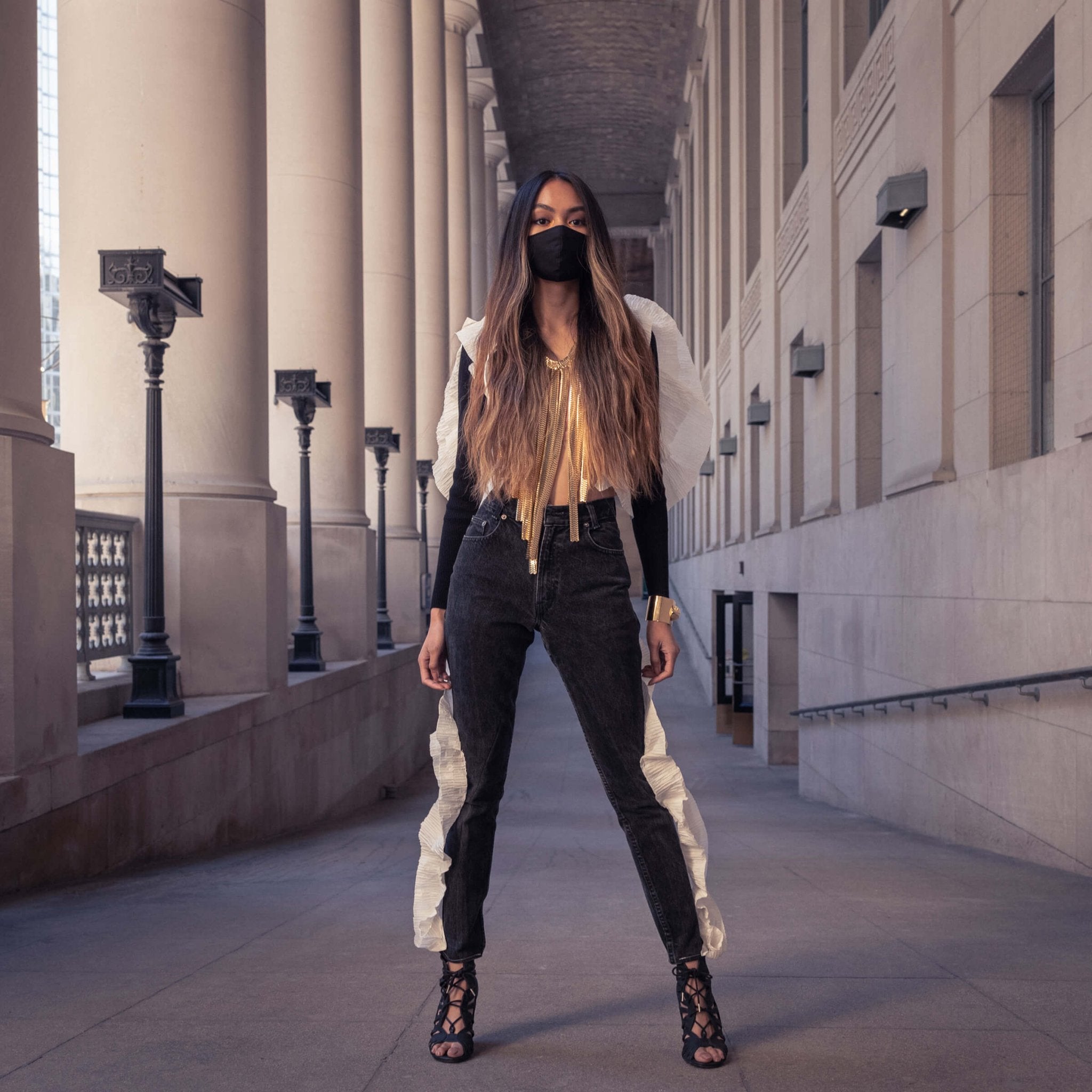 Striking model with wavy long ombré hair standing very confidently in an outdoor grand neoclassical colonnade.. She is wearing repurposed black jeans and a black bolero sweater with pleated paper ruffles sewn on to the clothes. The paper looks like wings on her back. Accented the look with a large gold fringe necklace for a dramatic look.