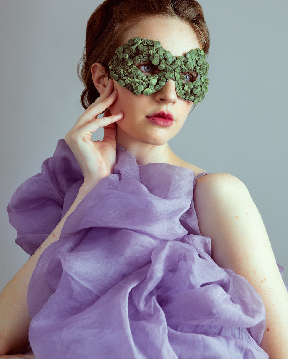 A model elegantly poses as she looks to the side gazing into the distance. Her auburn hair is tied back, and she wears a unique mask made from small round green leaves. She is wearing a frothy lavender organza gown that is draped diagonally across her body, while her right hand gently caresses her cheek.