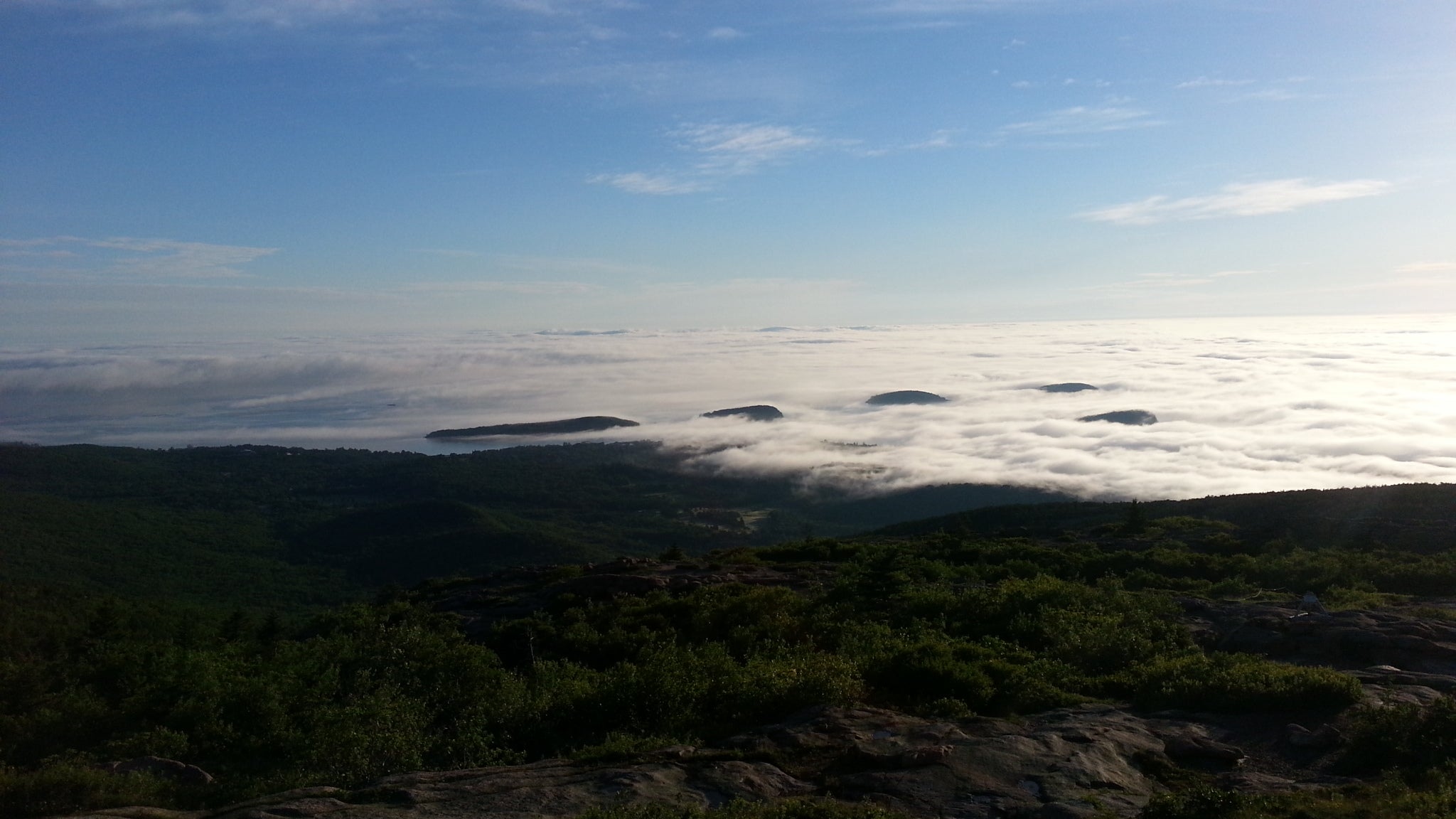 A panoramic view from a mountain top, showcasing rolling hills enveloped by a sea of thig fluffy clouds. The sky is bright blue with the sun having just risen. The landscape is lush with greenery, reflecting the beauty of nature and Demascaré's commitment to preserving it.