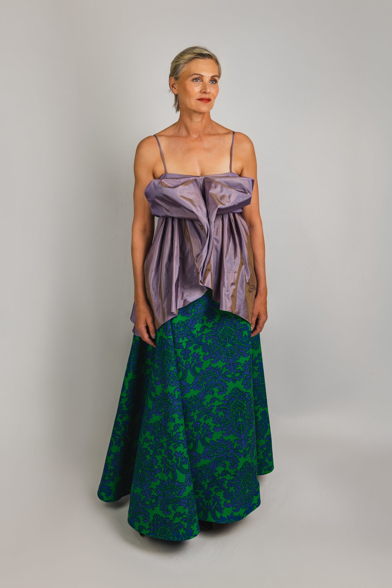 A blonde mature model wearing a luscious lavender satin bow-front top over a green and blue floral patterned  A-line skirt, photographed against a neutral light gray studio background.